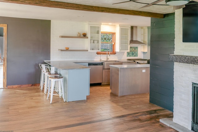 kitchen with wall chimney range hood, a sink, light wood-style floors, dishwasher, and a peninsula
