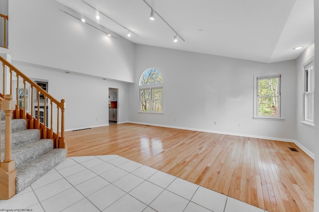 unfurnished living room with track lighting, stairs, baseboards, high vaulted ceiling, and light wood-style flooring