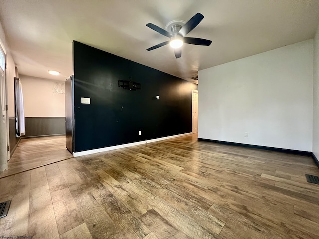 empty room with wood-type flooring, a wainscoted wall, a ceiling fan, baseboards, and visible vents
