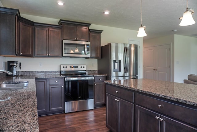 kitchen with appliances with stainless steel finishes, a sink, dark brown cabinetry, dark wood finished floors, and dark stone countertops