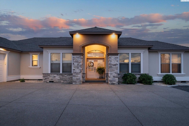 exterior entry at dusk with roof with shingles, stucco siding, stone siding, and crawl space