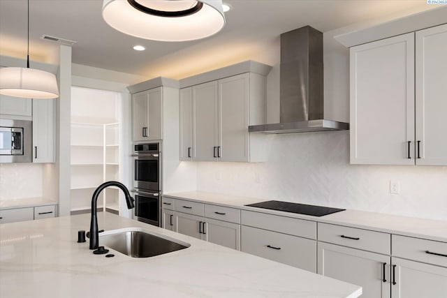kitchen with wall chimney range hood, light stone counters, a sink, stainless steel appliances, and decorative light fixtures