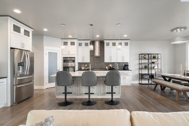 kitchen featuring pendant lighting, wall chimney range hood, appliances with stainless steel finishes, white cabinets, and a center island with sink