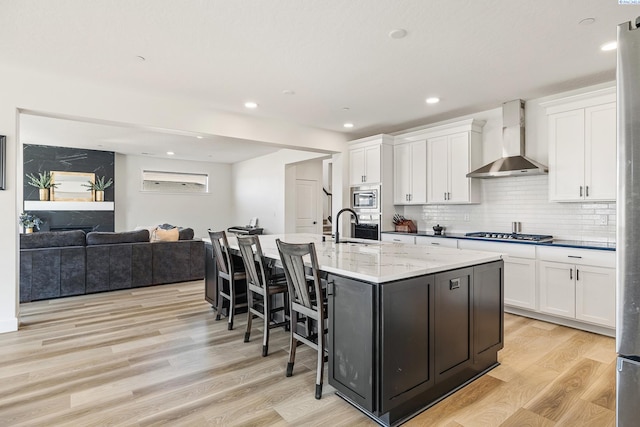 kitchen with white cabinets, wall chimney range hood, a center island with sink, and stainless steel appliances