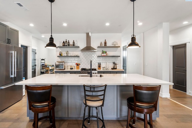 kitchen featuring high end refrigerator, light countertops, wall chimney exhaust hood, open shelves, and a large island with sink