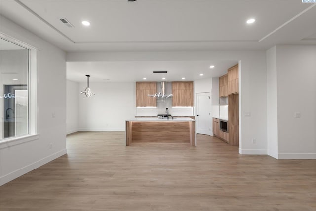 kitchen featuring tasteful backsplash, decorative light fixtures, light hardwood / wood-style flooring, an island with sink, and wall chimney range hood