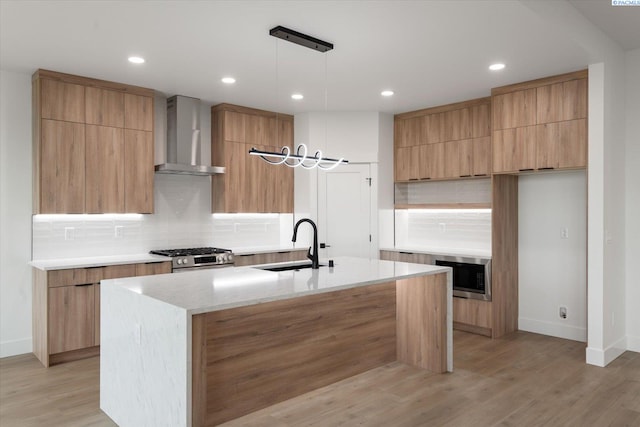 kitchen with appliances with stainless steel finishes, pendant lighting, sink, a center island with sink, and wall chimney exhaust hood