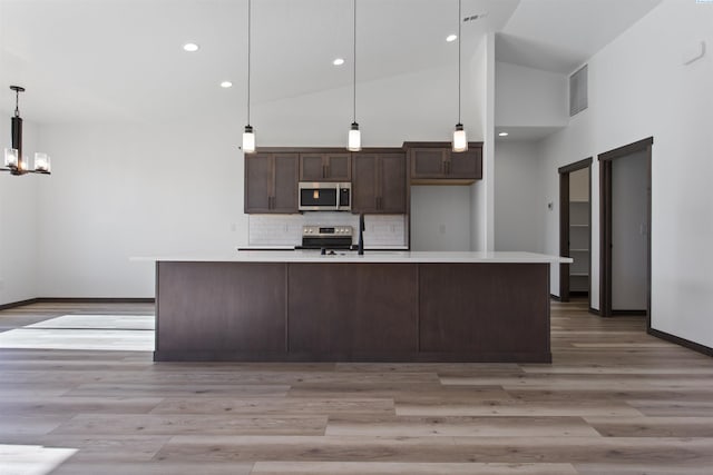 kitchen with appliances with stainless steel finishes, hanging light fixtures, backsplash, dark brown cabinets, and a center island with sink