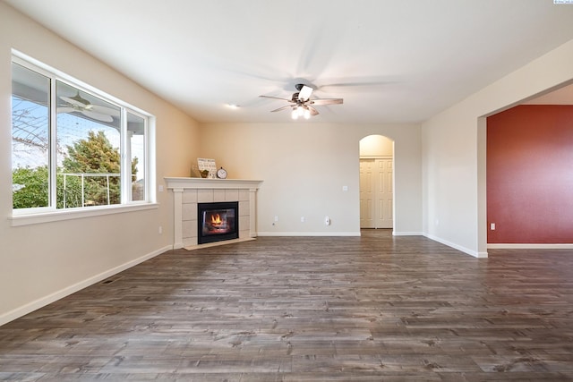 unfurnished living room featuring arched walkways, dark wood finished floors, ceiling fan, a tile fireplace, and baseboards