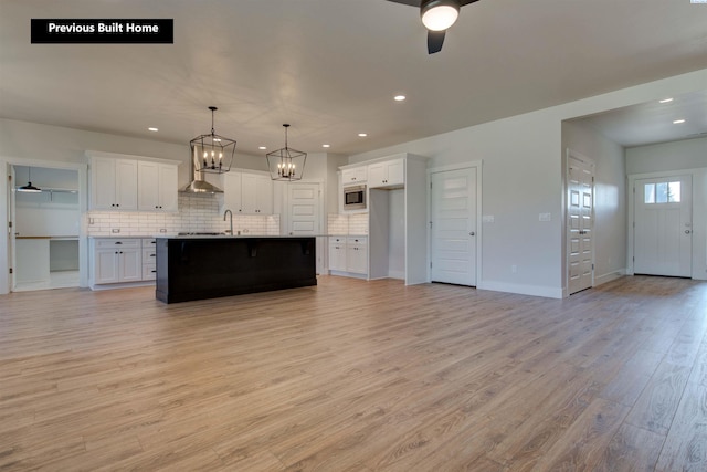 kitchen with ceiling fan with notable chandelier, wall chimney range hood, light wood-type flooring, tasteful backsplash, and stainless steel microwave