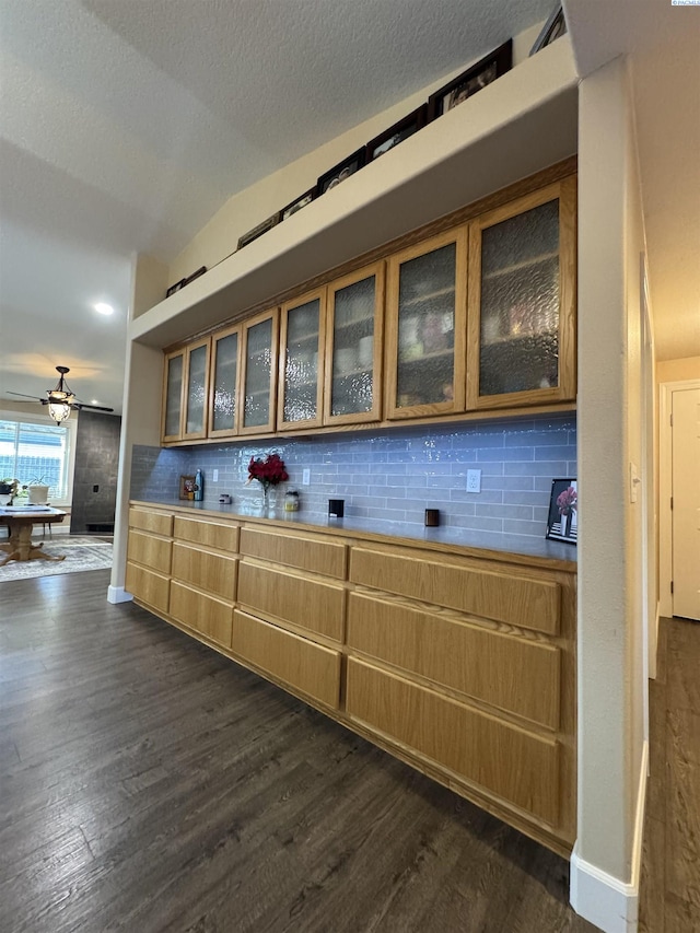 kitchen featuring backsplash, glass insert cabinets, vaulted ceiling, brown cabinetry, and dark wood-style flooring