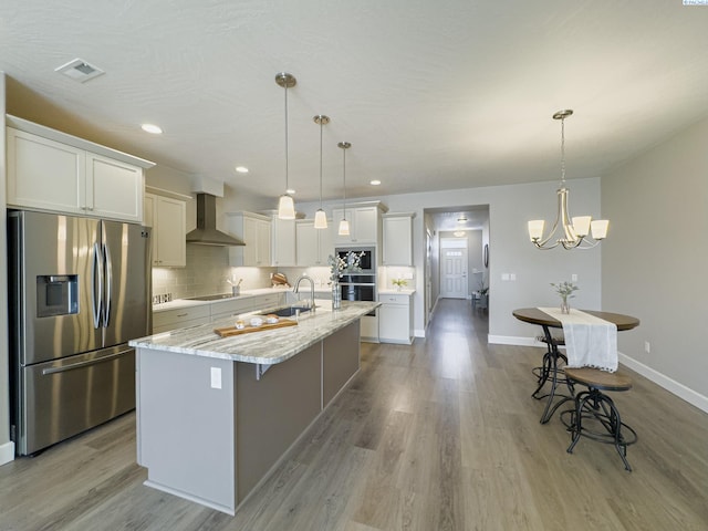 kitchen with light wood finished floors, a center island with sink, appliances with stainless steel finishes, wall chimney range hood, and a sink