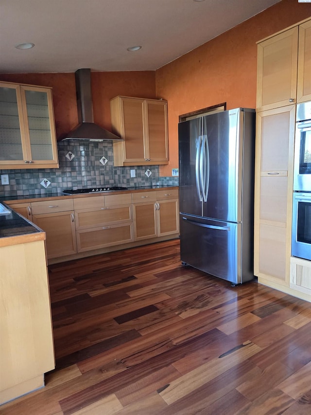 kitchen featuring dark wood-type flooring, appliances with stainless steel finishes, wall chimney range hood, tasteful backsplash, and dark countertops