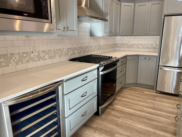 kitchen featuring gray cabinetry, wall chimney range hood, beverage cooler, appliances with stainless steel finishes, and light wood-style floors