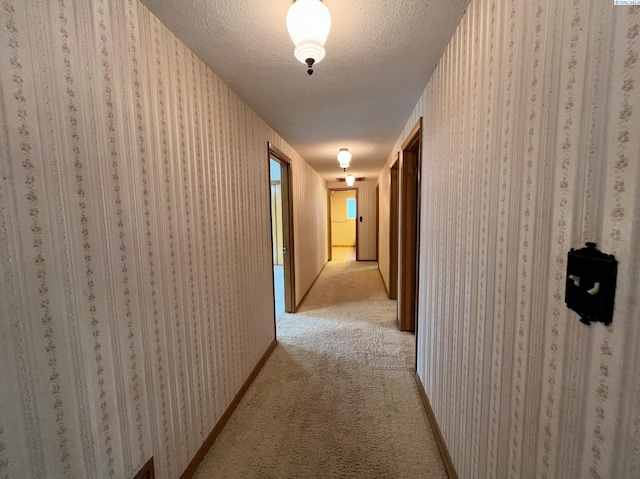 hallway with light colored carpet, a textured ceiling, baseboards, and wallpapered walls