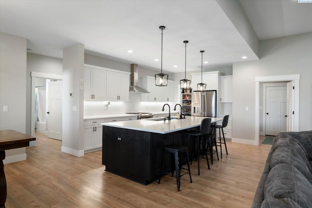 kitchen with high end fridge, hanging light fixtures, a center island with sink, wall chimney range hood, and white cabinets