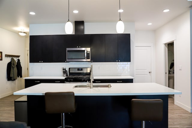 kitchen featuring tasteful backsplash, an island with sink, hanging light fixtures, stainless steel appliances, and light wood-type flooring