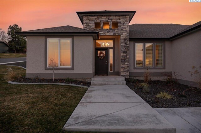 exterior entry at dusk featuring stone siding, a shingled roof, a yard, and stucco siding