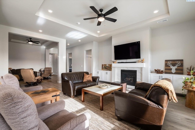 living room featuring a tiled fireplace, light hardwood / wood-style flooring, ceiling fan, and a tray ceiling