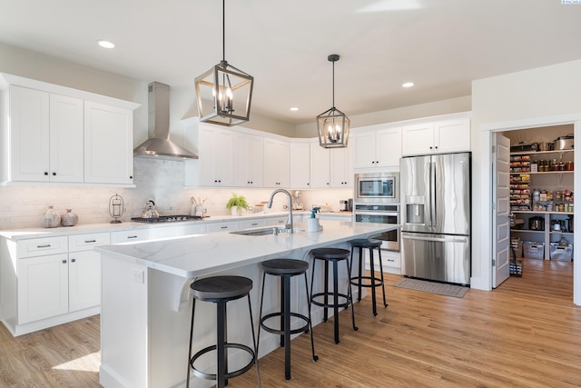 kitchen with an island with sink, wall chimney exhaust hood, light wood-style flooring, appliances with stainless steel finishes, and a sink