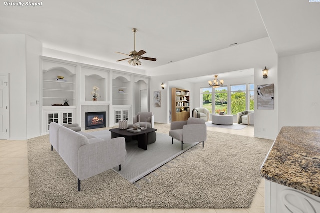 living room featuring ceiling fan with notable chandelier, built in features, a tiled fireplace, and light tile patterned flooring