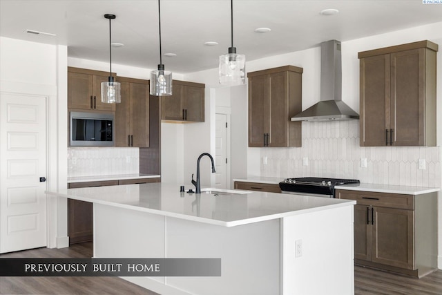 kitchen with wall chimney range hood, sink, hanging light fixtures, an island with sink, and built in microwave