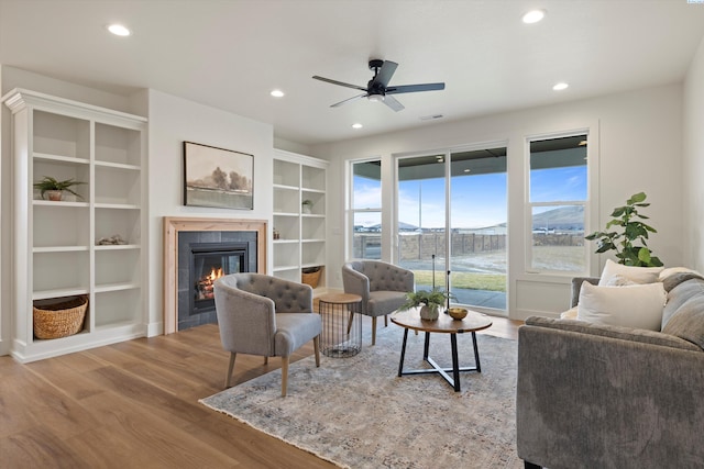 living room featuring built in features, hardwood / wood-style flooring, a tiled fireplace, ceiling fan, and a mountain view