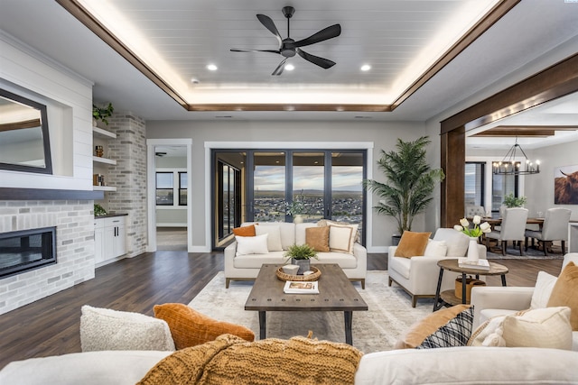 living room featuring dark wood-type flooring, a tray ceiling, ceiling fan with notable chandelier, and a brick fireplace