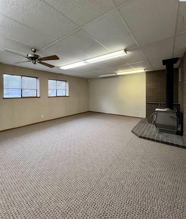 basement with a drop ceiling, carpet, ceiling fan, and a wood stove