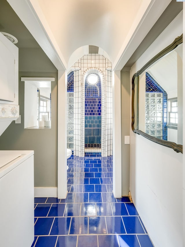 hallway with stacked washer / dryer, vaulted ceiling, a healthy amount of sunlight, and dark tile patterned flooring