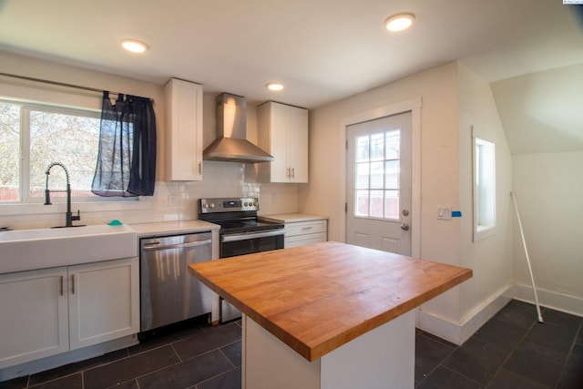 kitchen featuring wall chimney range hood, white cabinets, appliances with stainless steel finishes, a sink, and butcher block countertops