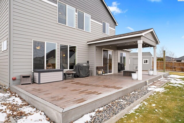 snow covered deck featuring an outbuilding, a fire pit, area for grilling, fence, and a storage unit