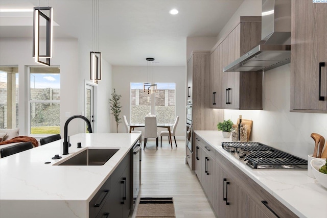 kitchen with sink, stainless steel appliances, an island with sink, decorative light fixtures, and wall chimney exhaust hood