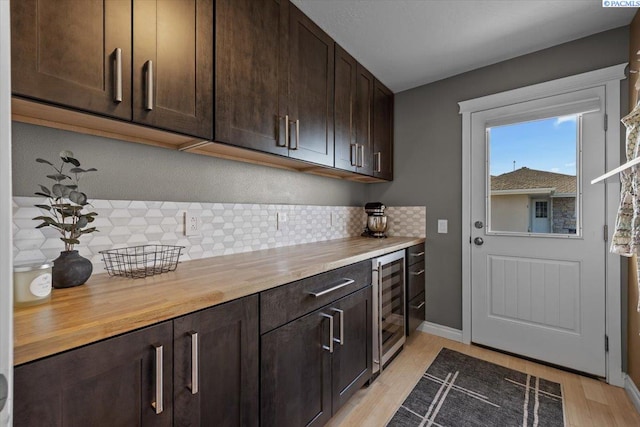 kitchen featuring dark brown cabinetry, beverage cooler, tasteful backsplash, light wood-style flooring, and wood counters