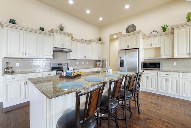 kitchen featuring sink, stainless steel appliances, light stone counters, an island with sink, and dark hardwood / wood-style flooring