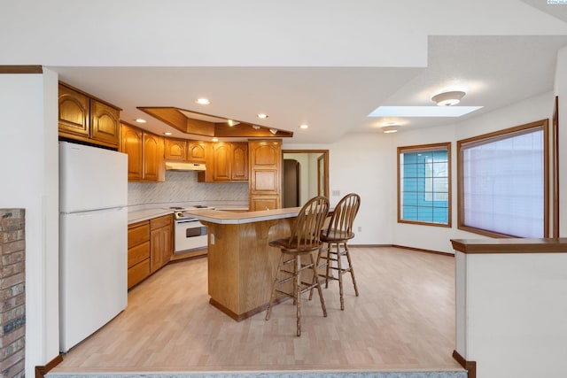 kitchen with a breakfast bar, brown cabinets, light wood-style flooring, white appliances, and under cabinet range hood