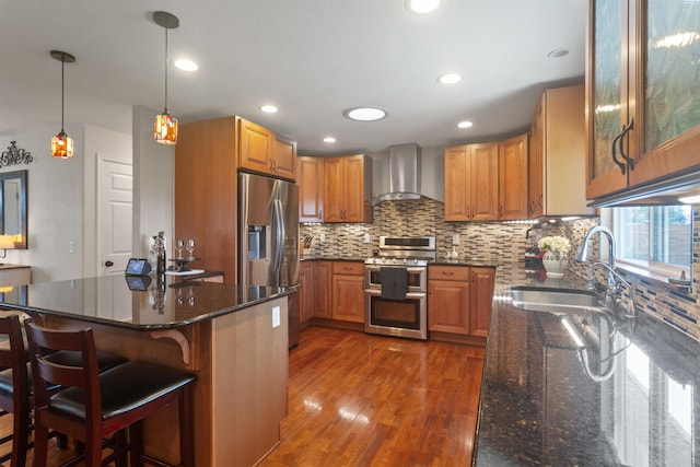 kitchen featuring a sink, wall chimney exhaust hood, dark stone countertops, stainless steel appliances, and dark wood-type flooring