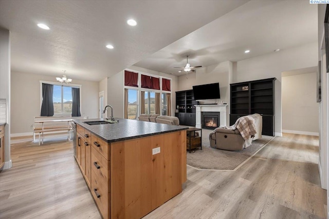 kitchen with light wood finished floors, dark countertops, a glass covered fireplace, a sink, and ceiling fan with notable chandelier