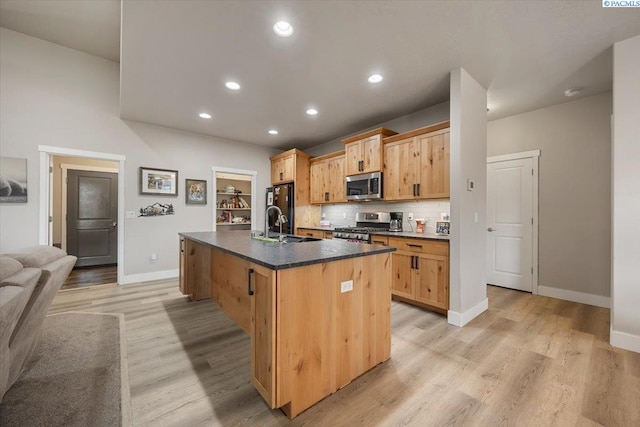 kitchen with tasteful backsplash, dark countertops, appliances with stainless steel finishes, a kitchen island with sink, and light wood-style floors