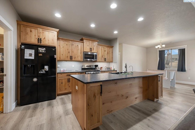 kitchen featuring dark countertops, light wood-style flooring, decorative backsplash, appliances with stainless steel finishes, and an island with sink