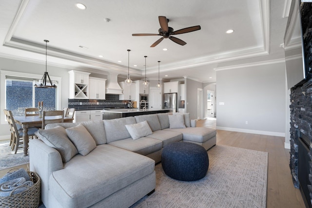 living room with a raised ceiling, light wood-style floors, ornamental molding, baseboards, and ceiling fan with notable chandelier