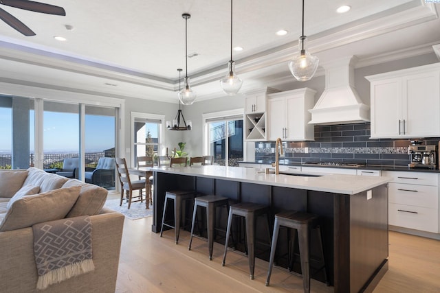 kitchen featuring a tray ceiling, light wood finished floors, custom exhaust hood, open floor plan, and a sink