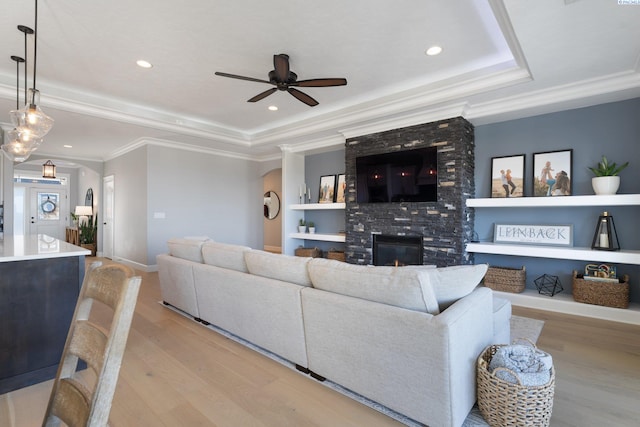 living area featuring crown molding, a fireplace, a raised ceiling, a ceiling fan, and light wood-type flooring