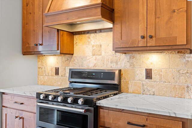 kitchen with premium range hood, decorative backsplash, brown cabinets, light stone countertops, and gas range