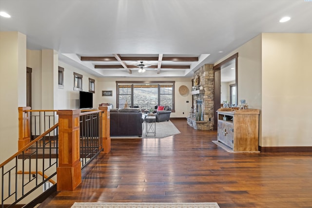 living room with dark wood-type flooring, coffered ceiling, a stone fireplace, beamed ceiling, and ceiling fan