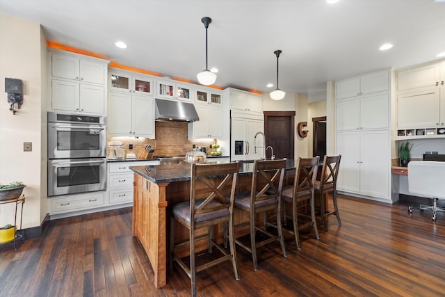 kitchen featuring double oven, hanging light fixtures, a center island with sink, a kitchen bar, and dark hardwood / wood-style flooring