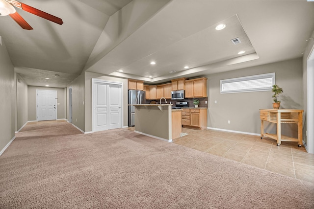 kitchen featuring appliances with stainless steel finishes, a kitchen breakfast bar, a tray ceiling, an island with sink, and light carpet