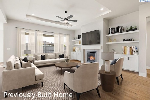 living room featuring ceiling fan, a tray ceiling, a tile fireplace, and light hardwood / wood-style flooring