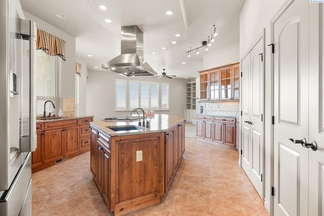 kitchen featuring decorative backsplash, island range hood, a center island with sink, brown cabinets, and a ceiling fan