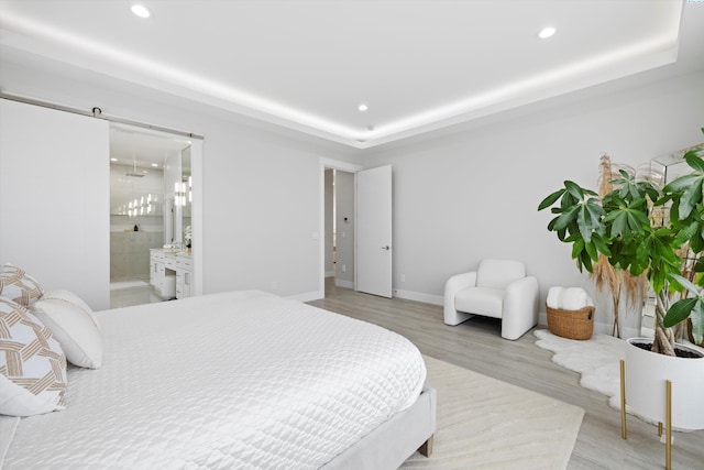 bedroom featuring ensuite bath, a barn door, a raised ceiling, and light hardwood / wood-style floors